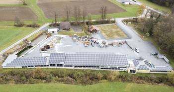 Eine Luftaufnahme eines Gebäudes mit Solarpanelen auf dem Dach. Das Gebäude ist von einem Parkplatz und einer Straße umgeben. In der Nähe befinden sich Bäume, ein Feld und ein Haus.