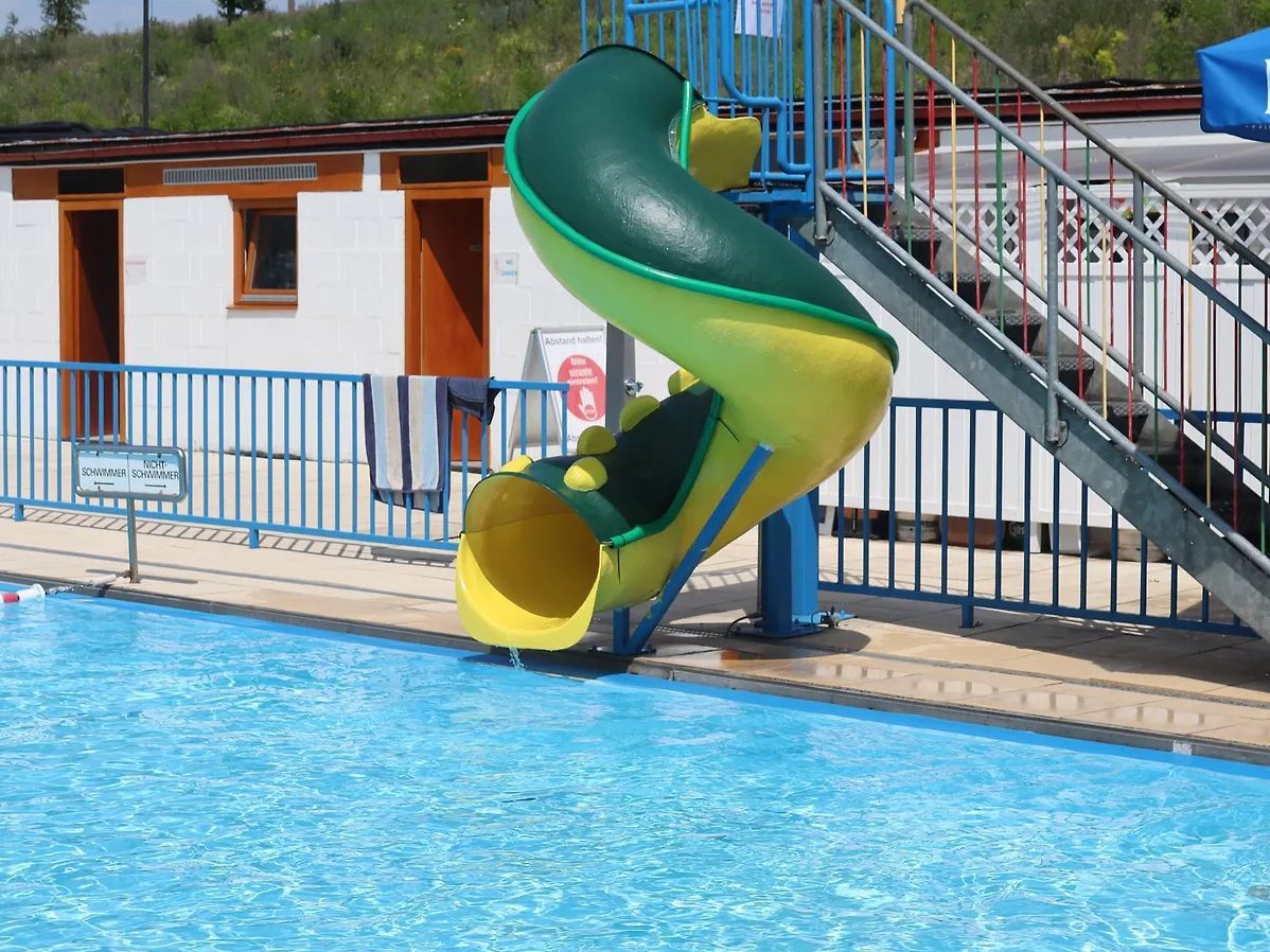 Außenschwimmbad mit blauer Umzäunung, blauem und gelbem Wasserrutsche und blauem Dach an einem sonnigen Tag.