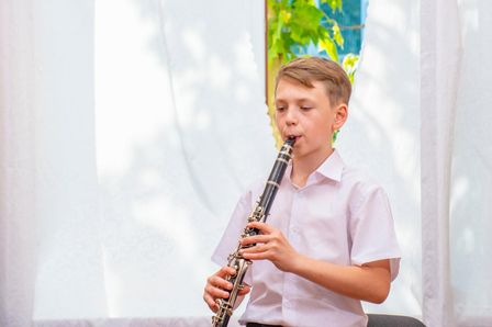 Ein junger Junge spielt Klarinette, während er in einem Raum mit einem weißen Vorhang im Hintergrund sitzt.