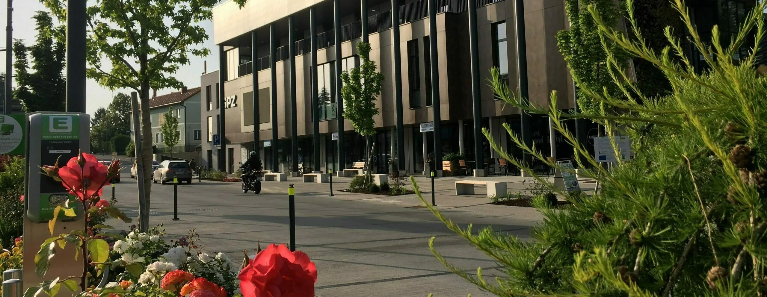 Ein modernes Gebäude mit vielen Fenstern und Balkonen. Eine Person auf einem Motorrad ist davor. Es gibt Bänke und Pflanzen im Bereich.