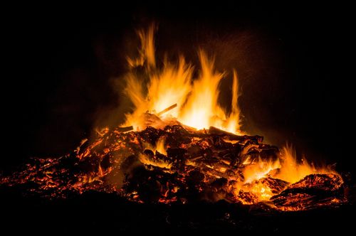 Ein Lagerfeuer brennt nachts hell, die Flammen reichen bis in den dunklen Himmel. Das Feuer ist von Holz umgeben, einiges noch intakt, anderes brennt.