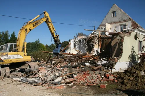 Ein gelber Bagger bricht ein Haus ab. Das Haus ist in Trümmern, mit herumliegenden Trümmern. Bäume sind im Hintergrund sichtbar.