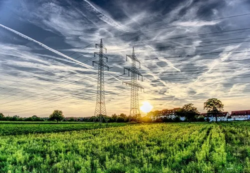 Sonnenuntergang über einem Feld mit zwei hohen Stromleitungsmasten im Vordergrund. Der Himmel ist bewölkt und die Sonne geht hinter den Bäumen unter.