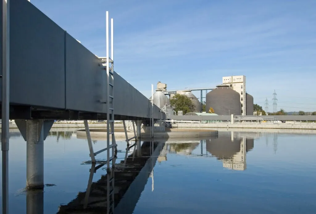 Eine Wasseraufbereitungsanlage mit einem großen, gebogenen Tank und einer Brücke, die sich über einen Wasserkörper erstreckt, wobei sich der Himmel und die umliegenden Strukturen im Wasser spiegeln.