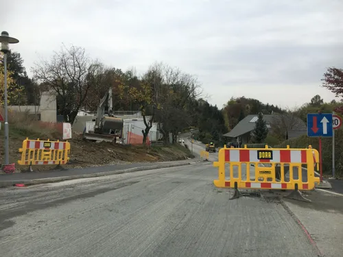 Eine Straße im Bau mit einem schweren Baufahrzeug auf der Seite, Absperrungen an den Seiten und Häusern und Bäumen im Hintergrund.