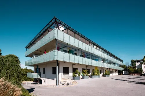 Ein mehrstöckiges Gebäude mit Glasbalkonen, umgeben von Pflanzen und Bäumen, steht unter einem klaren blauen Himmel.