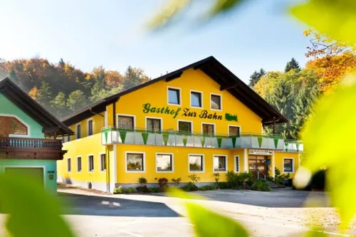 Ein leuchtend gelbes Gebäude mit einem Schild, das Gasthof Zur Bahn liest, ist von üppigem Baumbestand und Grün umgeben. Das Gebäude hat mehrere Fenster und einen Balkon mit Zaun.