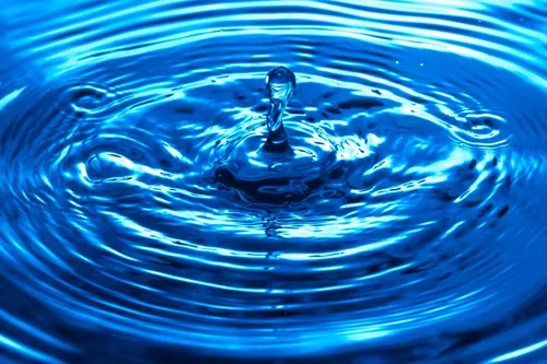 Ein Wassertropfen fällt in einen blauen Wasserkörper und erzeugt konzentrische Wellen. Das Wasser ist klar und hat einen blauen Farbton.