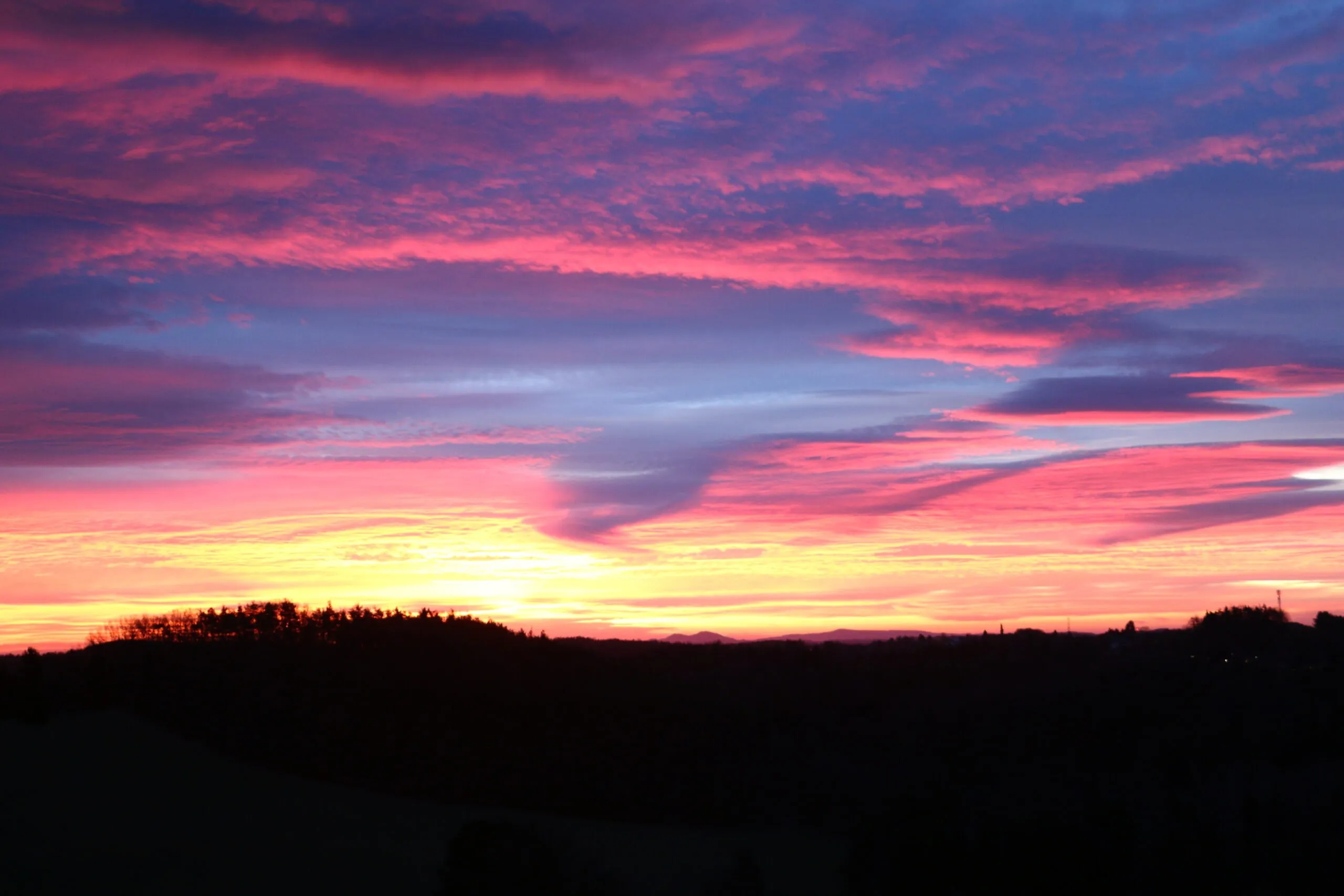 Sonnenuntergang über einer hügeligen Landschaft mit lebhaften Farben von Rosa, Lila und Gelb am Himmel. Bäume sind vor dem farbenfrohen Horizont silhouettiert.