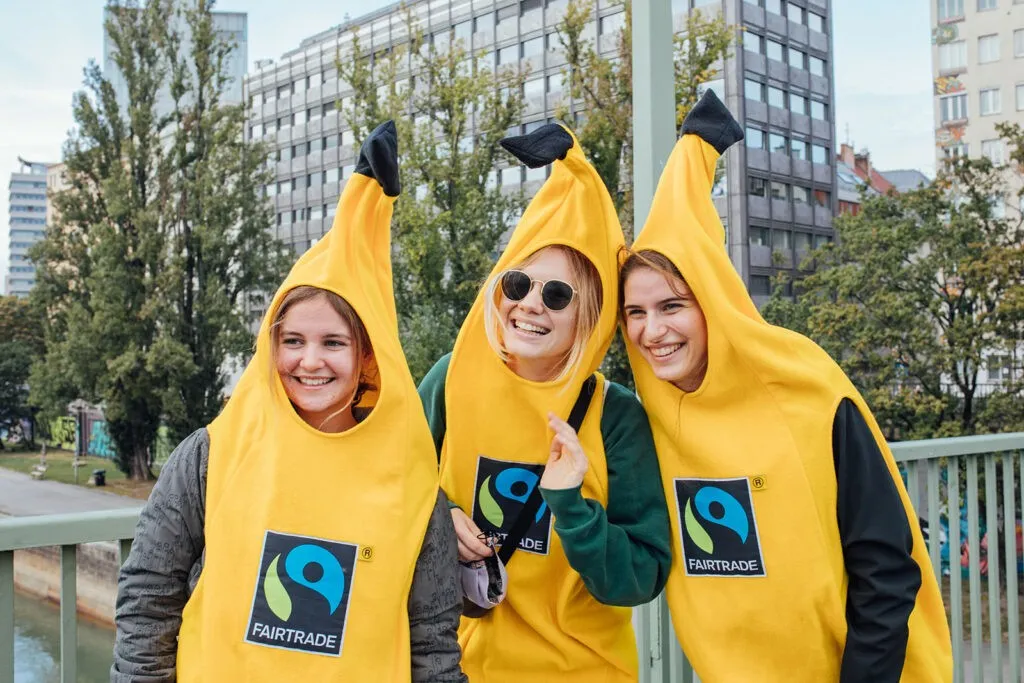 Drei Frauen in Bananenkostümen posieren für ein Foto vor einem Gebäude mit großen Fenstern. Die Kostüme tragen ein Fairtrade-Logo.