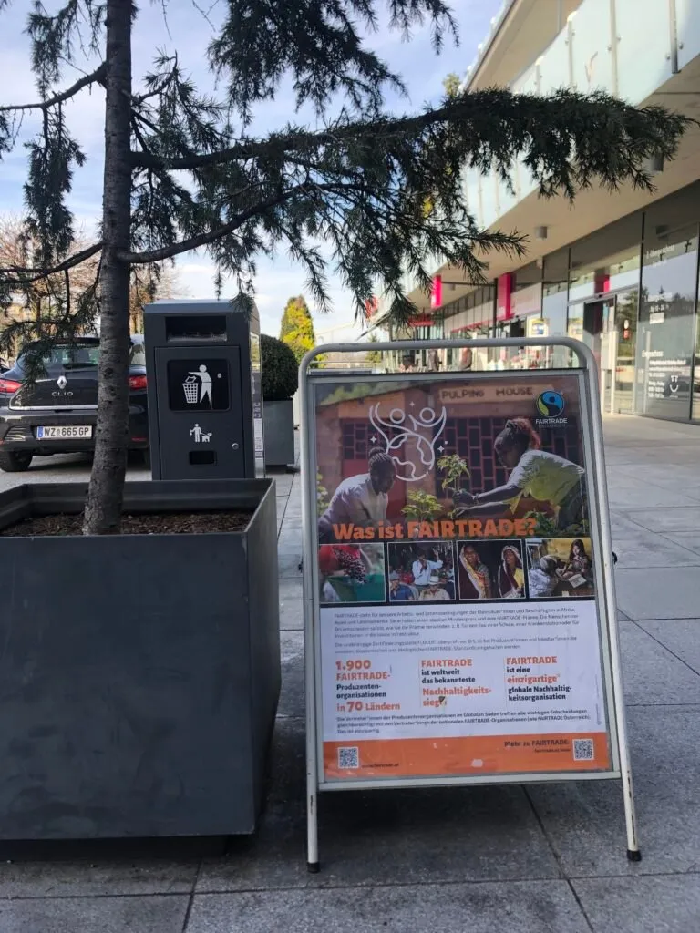 Ein Poster über Fairtrade steht neben einem Mülleimer in einer Außenumgebung. Ein Auto ist hinter dem Mülleimer geparkt. Das Poster zeigt arbeitende Menschen und erklärt das Konzept von Fairtrade.