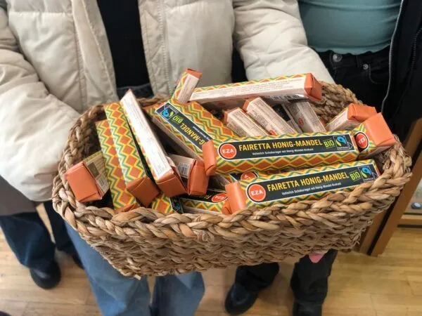 Eine Person hält einen Korb voller orangefarbener Produkte mit der Aufschrift 'Fairetta Honig-Mandel Bio'. Der Korb ist aus gewebtem Material und die Person steht auf einem Holzboden.