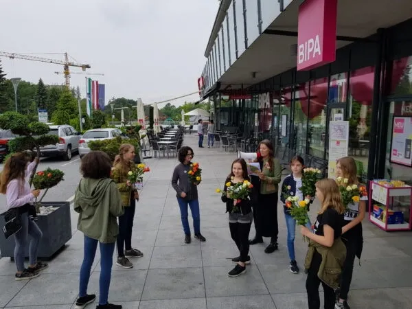 Eine Gruppe junger Frauen mit Blumen steht vor einem Gebäude mit einem BIPA-Schild. Im Hintergrund befinden sich Bäume und geparkte Autos.