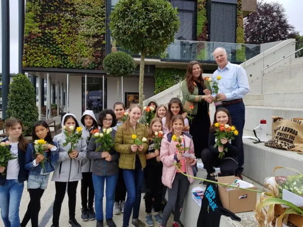 Eine Gruppe junger Mädchen und zwei Erwachsene halten Blumen vor einem Gebäude mit einer grünen Wand und Treppen.