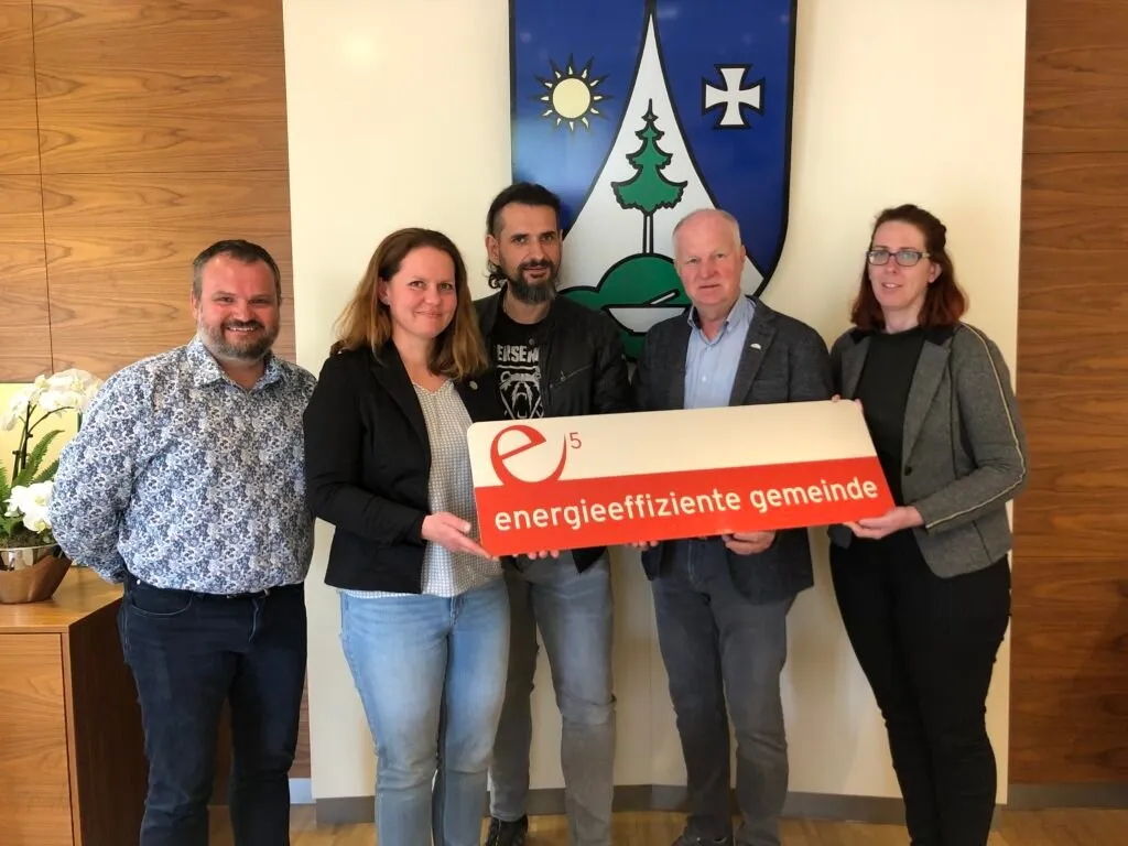Fünf Personen stehen lächelnd zusammen in einem Raum. Sie halten eine weiße Box mit rotem Text, der 'energieeffiziente gemeinde' lautet. Hinter ihnen befindet sich ein blau-weißes Banner mit einem Kreuz und einer Sonne.