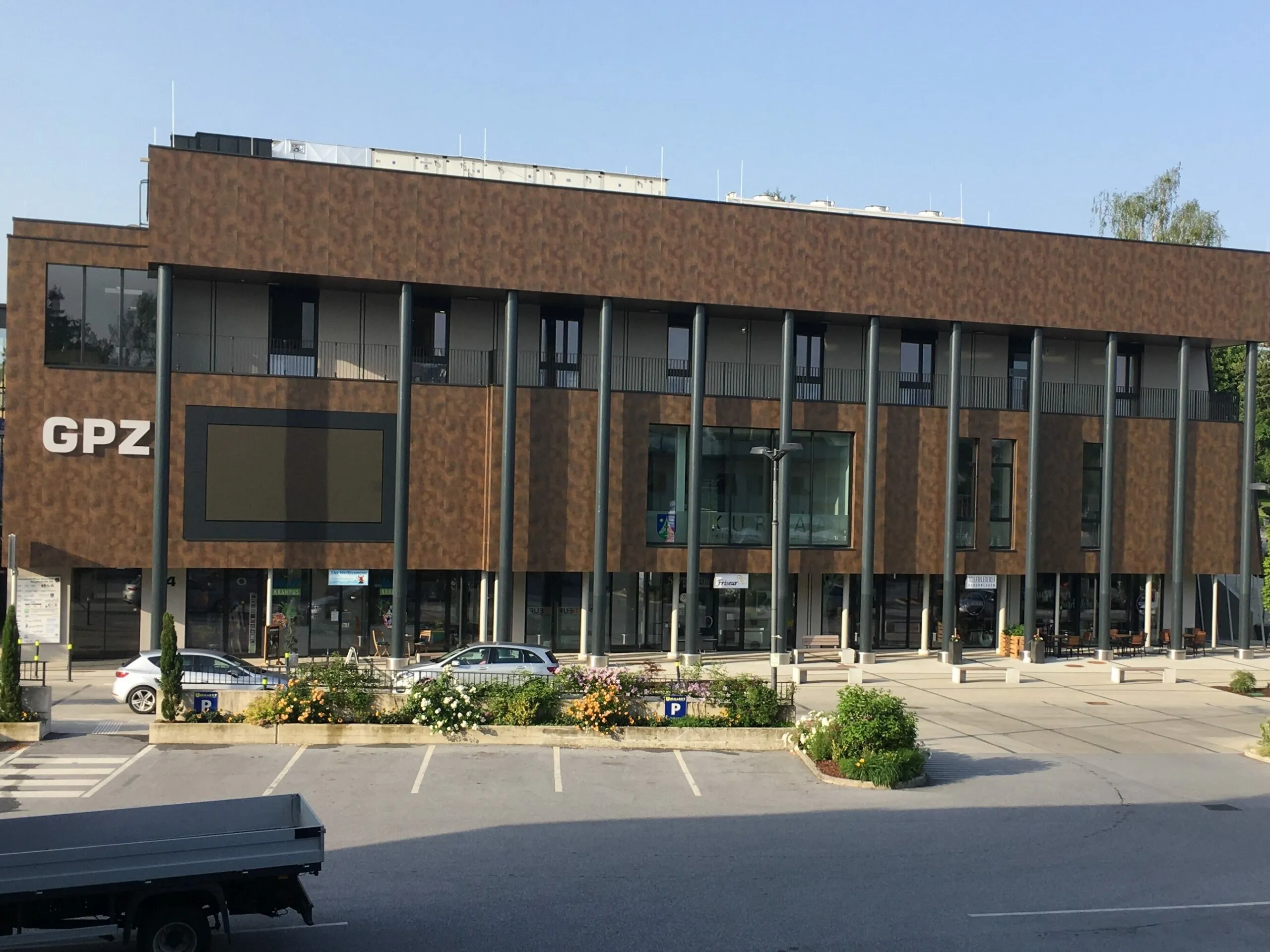 Ein modernes Gebäude mit brauner Fassade, mehreren Stockwerken und Glasfenstern. Vor dem Gebäude geparkte Autos. Großer Parkplatz mit mehreren Autos. Bäume und Pflanzen.