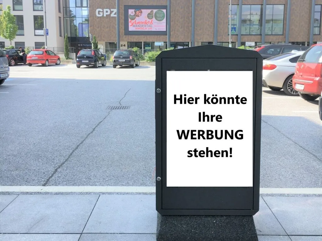 Ein Schild am Bürgersteig lädt Unternehmen ein, hier ihre Werbung zu platzieren. Hinter dem Schild sind Autos vor einem Gebäude mit einer großen digitalen Anzeige geparkt.