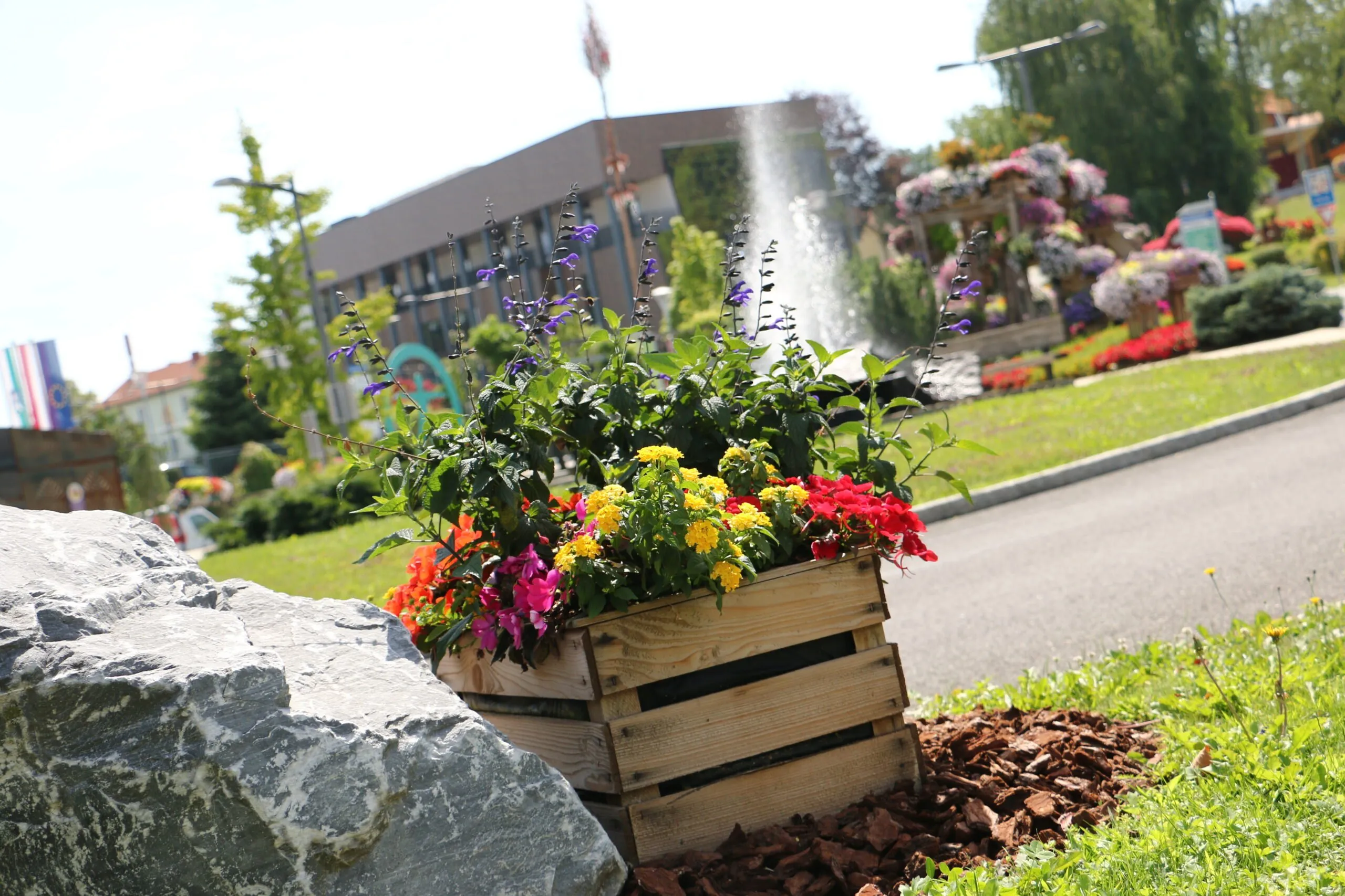 Ein hölzerner Blumenkasten mit bunten Blumen steht am Straßenrand neben einem großen Stein, mit einem Gebäude und einem Brunnen im Hintergrund.