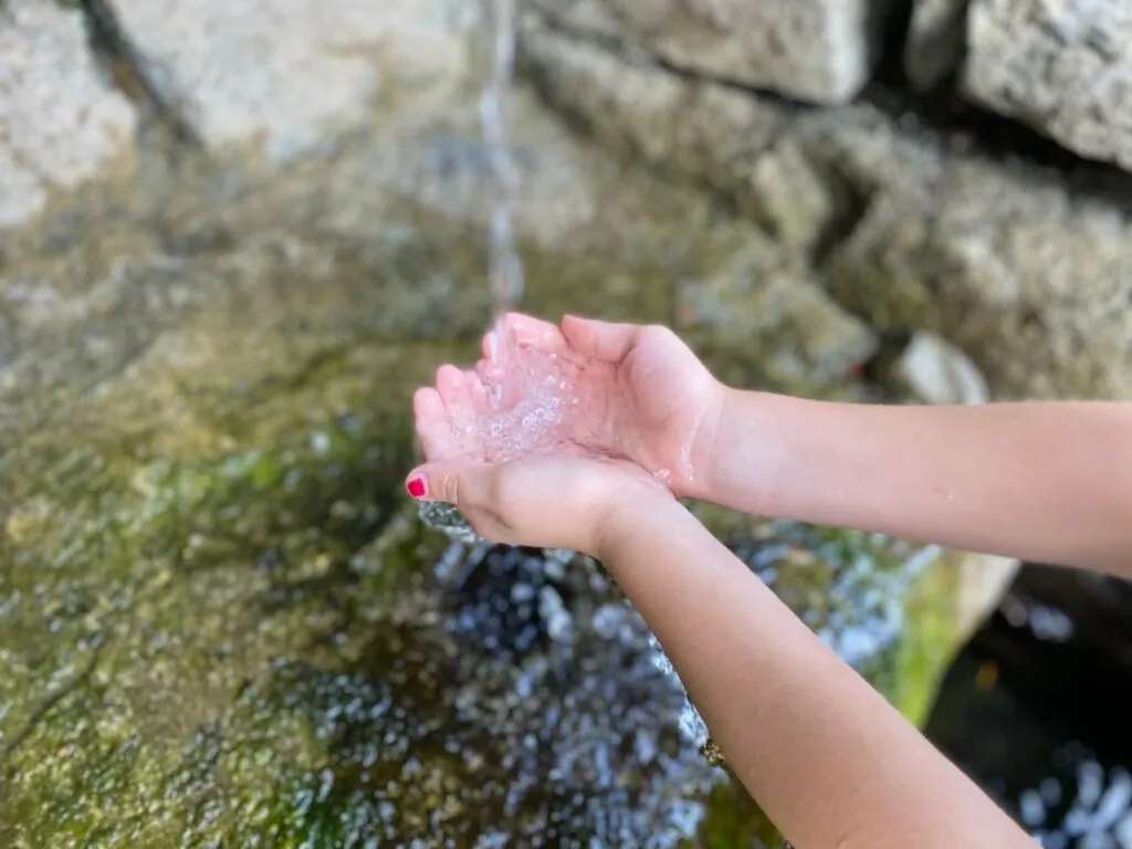 Ein Nahaufnahme der Hände eines Kindes, das Wasser aus einem Wasserfall auffängt, mit Wassertropfen auf den Handflächen und Fingern.
