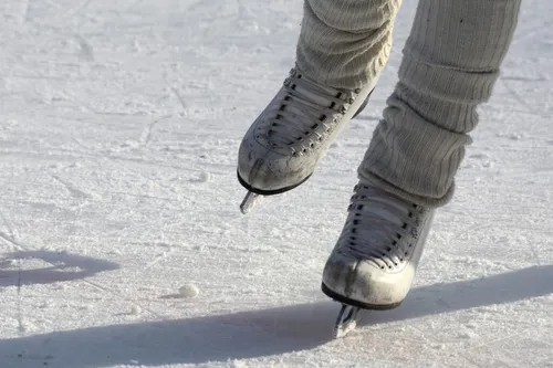 Nahaufnahme der Beine einer Person, die weiße Eislaufschuhe auf einer Eisbahn trägt, mit sichtbaren Schlittschuhblättern und Schneespuren.