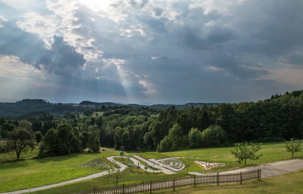 Luftaufnahme eines üppigen grünen Feldes mit einer Spirale auf dem Boden, einem Zaun und Bäumen. Der Himmel ist bewölkt, und Sonnenstrahlen dringen durch.