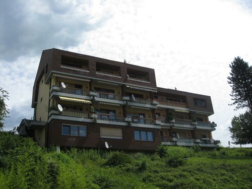 Ein mehrstöckiges Gebäude mit braunen Außenwänden und mehreren Balkonen, vor einem bewölkten Himmel. Das Gebäude ist von üppigem grünem Bewuchs umgeben.