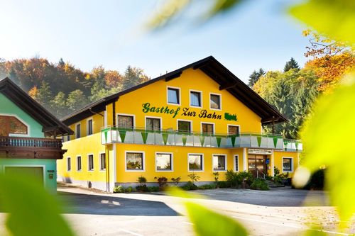 Ein hellgelbes Gebäude mit grünen Verzierungen namens Gasthof Zur Bahn steht vor einer Kulisse von Bäumen.