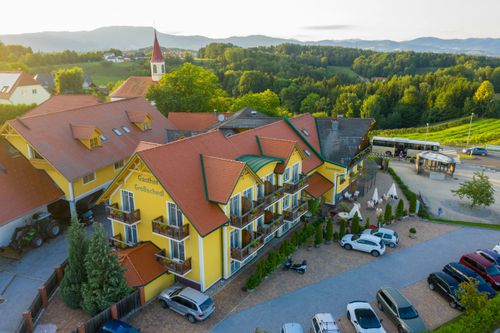 Ein Luftbild von Gasthof Großschedl, einem gelben Hotel mit rotem Dach, eingebettet in üppiger Grün- und Berglandschaft. Mehrere Autos sind vor dem Hotel geparkt, und ein Bus ist auf der Straße zu sehen.