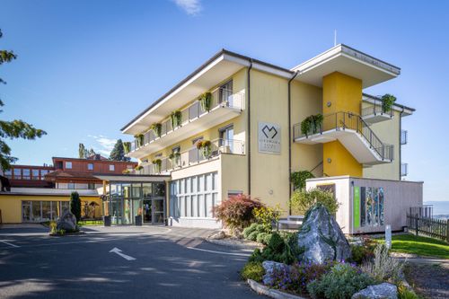 Ein modernes gelbes Hotel mit Balkonen, einem Logo und Treppen. Es ist von Pflanzen, Felsen und einem Schild umgeben. Der Himmel ist klar und blau.
