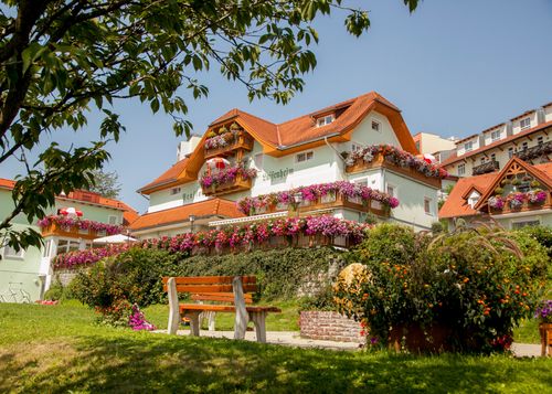 Ein großes, farbenfrohes Haus mit orangefarbenen Dächern und zahlreichen Blumenkästen ist zu sehen. Eine Holzbank steht im Vordergrund.