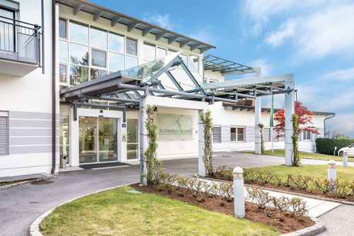 Der Eingang zur Schwarzel Klinik hat ein Glasdach, Glastüren und ein Schild. Das Gebäude hat viele Fenster und im Vordergrund gibt es Pflanzen und einen Laternenpfosten.