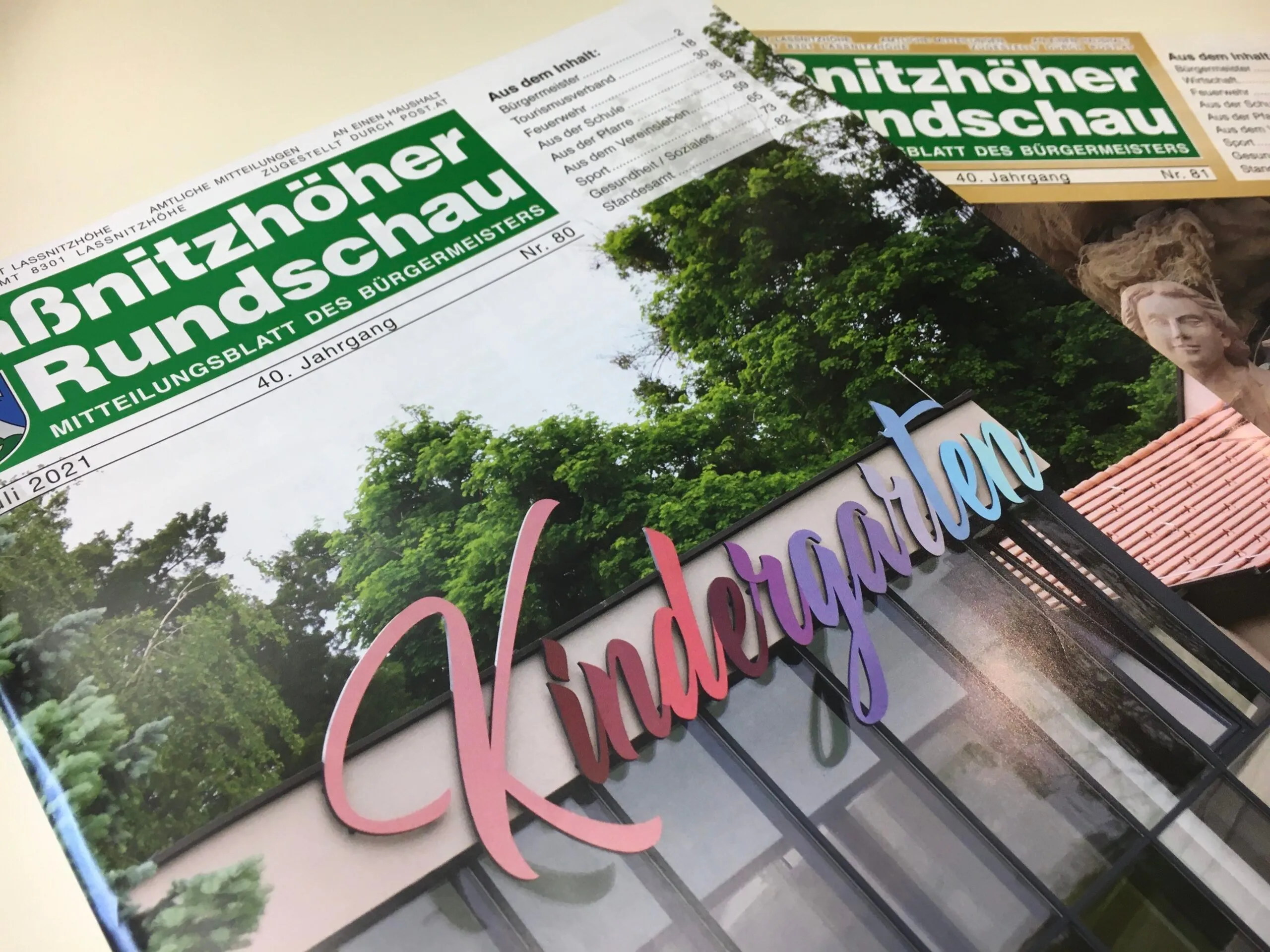 Nahaufnahme eines farbenfrohen Schildes mit der Aufschrift 'Kindergarten' auf einem Gebäude mit grüner Vegetation im Hintergrund. Das Bild zeigt auch ein grünes und weißes Dokument mit dem Titel 'Mittzhöher Landschau'.