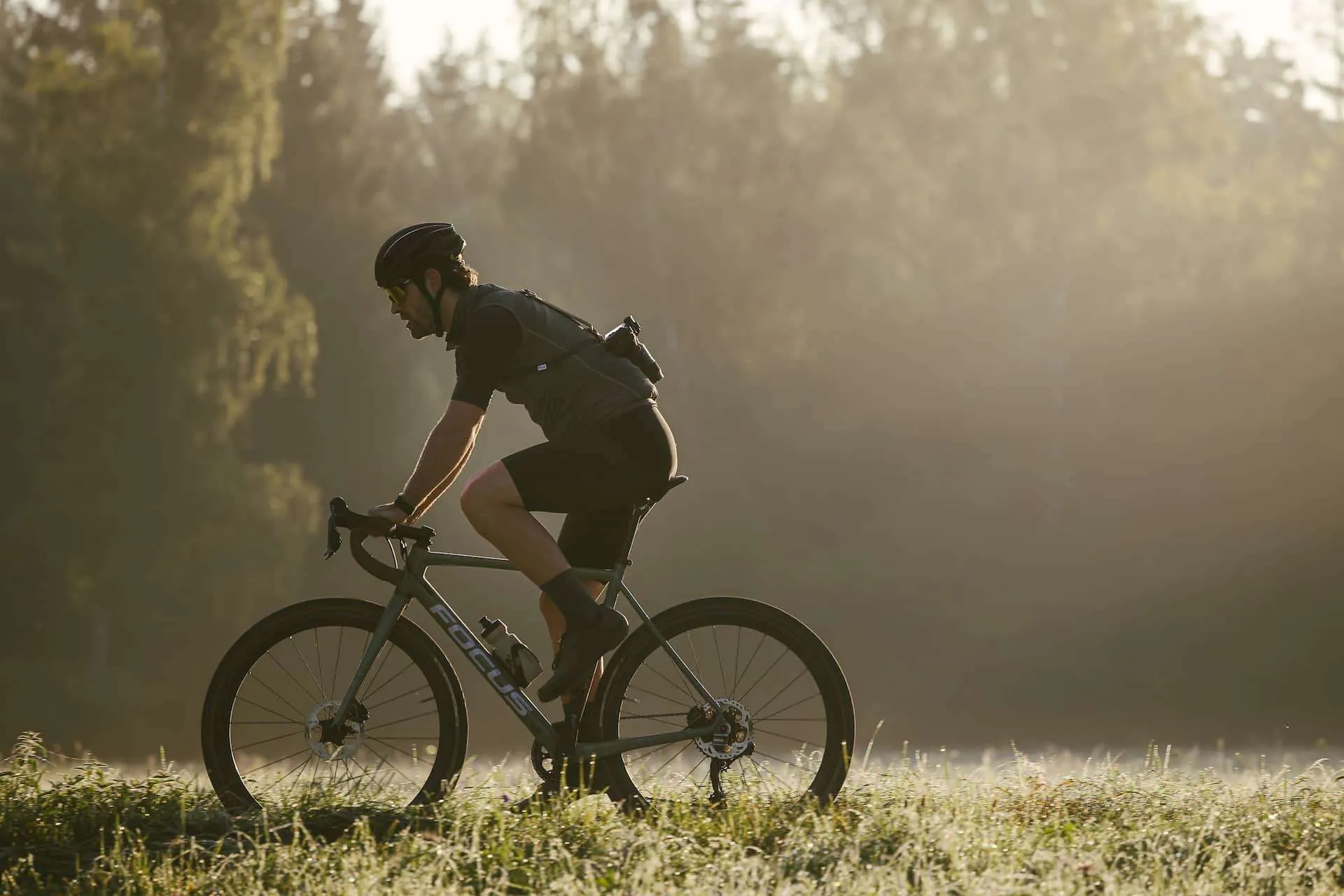 Ein Mann fährt ein Focus-Fahrrad auf einer Wiese mit Waldkulisse im Hintergrund. Er trägt einen Helm, Sonnenbrille und ein Radsporttrikot.