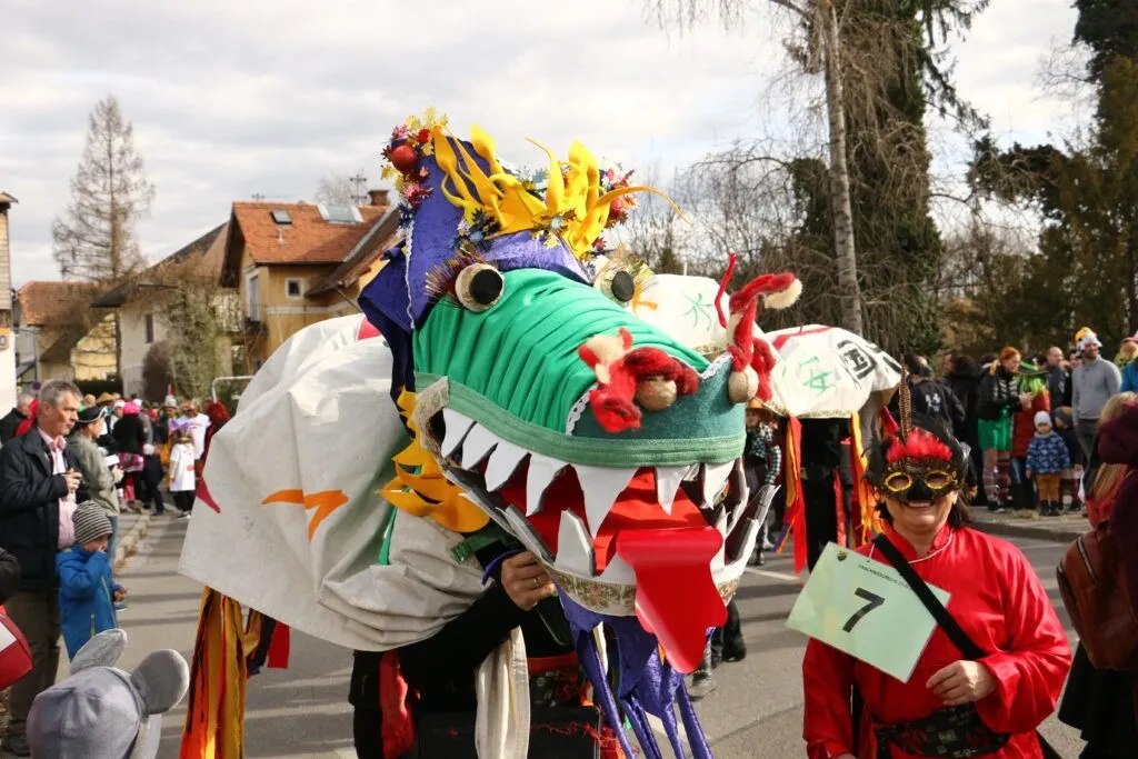 Eine Person mit einer Drachenmaske hält ein Schild mit der Nummer 7. Die Maske ist farbenfroh mit einem grünen Körper, roten Akzenten und einer gelb-lila Mütze. Dahinter laufen einige Leute auf der Straße.