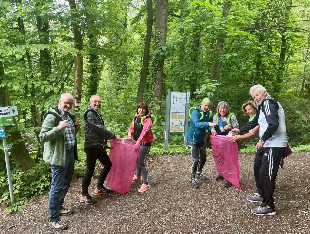 Eine Gruppe von Menschen im Wald, die rosa Beutel halten, lächeln und für ein Foto posieren. Hinter ihnen steht ein Schild mit Text und einem Bild.