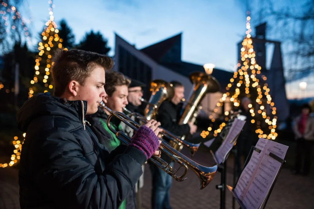 Eine Gruppe von Musikern spielt im Freien abends Blechblasinstrumente, umgeben von beleuchteten Weihnachtsbäumen.