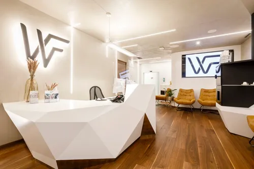 Ein moderner Empfangsbereich mit einem weißen Schreibtisch und zwei braunen Stühlen. An der Wand ist ein großer Monitor mit dem WF-Logo angebracht.