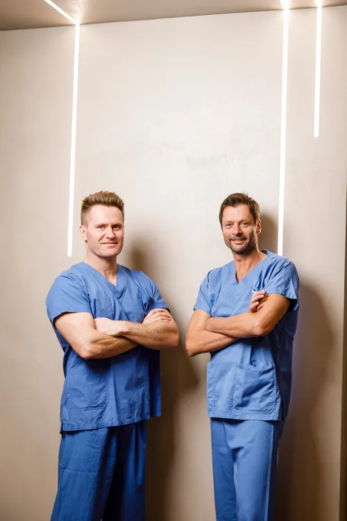 Zwei Männer in blauen Scrubs stehen mit verschränkten Armen vor einer weißen Wand mit weißen Linien. Sie scheinen sich in einem professionellen Umfeld zu befinden, möglicherweise in einem Krankenhaus.