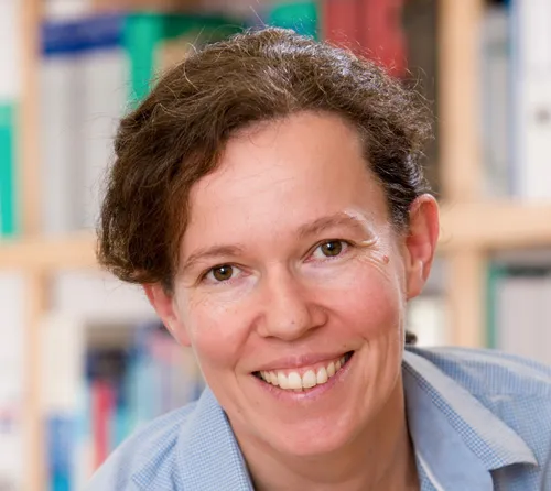 Eine lächelnde Frau mit kurzen braunen Haaren und einem blauen Hemd steht vor einem Bücherregal.