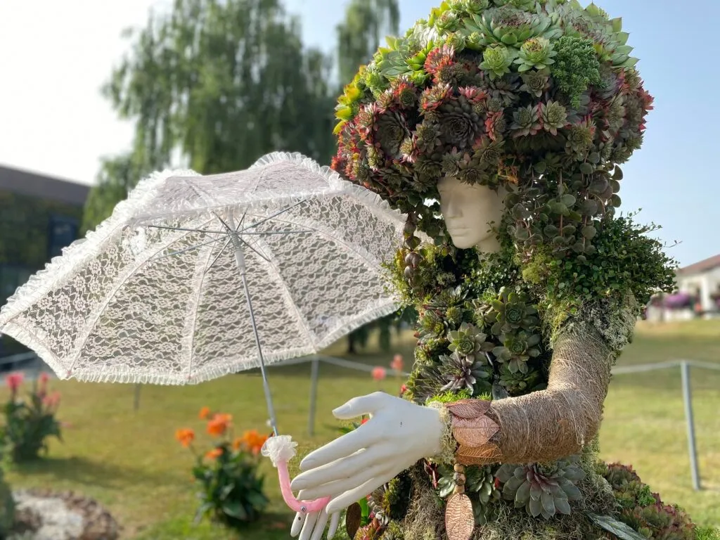 Eine Puppe im Garten mit einem Hut aus Sukkulenten, der eine rosa Spitzenschirm hält. Grüner Rasen und orange Blumen sind hinter der Puppe. Bäume sind im Hintergrund.