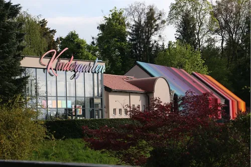 Ein Kindergartengebäude mit Glasfassade und buntem Schild, umgeben von Grün und Bäumen.