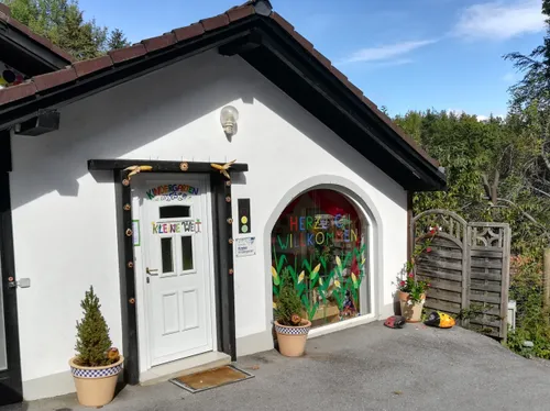 Ein kleines, weißes Gebäude dient als Kindergarten mit einem Glasfenster, das Herzlich Willkommen (Herzlich Willkommen) steht. Davor befinden sich Topfpflanzen und Dekorationen.