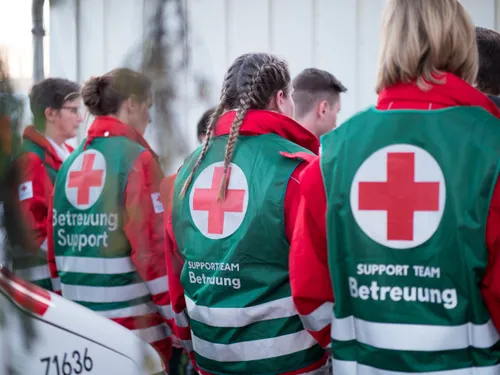 Freiwillige in grünen und roten Uniformen mit roten Kreuzen auf dem Rücken stehen in einer Reihe, möglicherweise in einem Gesundheitseinrichtung.