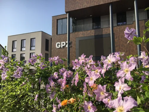 Ein Gebäude mit den Buchstaben GPZ an der Seite ist von einem Strauch mit lila Blumen umgeben. Dahinter stehen zwei Gebäude.