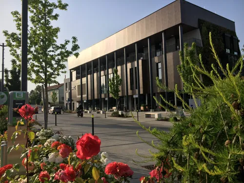 Ein modernes Gebäude mit grüner Fassade und großen Fenstern steht in einem Landschaftsbereich, vor dem ein Motorradfahrer vorbeifährt.