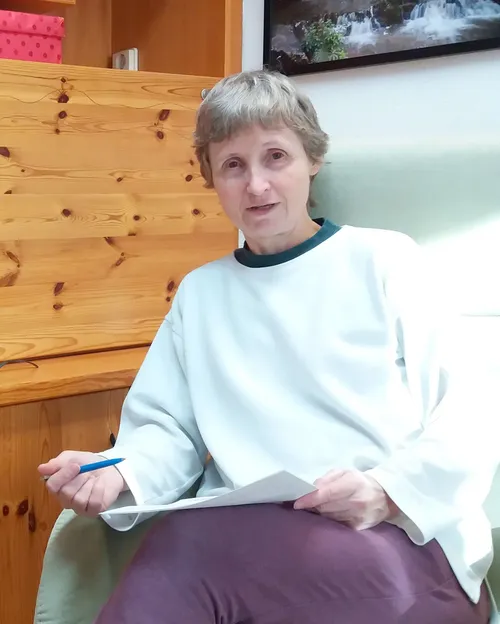 Eine ältere Frau in einem weißen Shirt sitzt auf einem Stuhl und hält einen Stift und ein Blatt Papier. Sie scheint konzentriert zu lesen oder zu schreiben. Hinter ihr ist eine Holzwand.