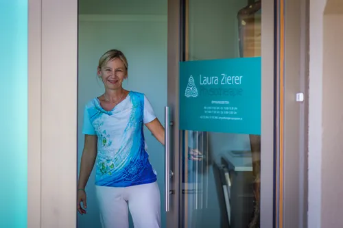 Eine Frau in einem blau-weißen Shirt steht vor einer Glastür mit einem Schild, das 'Laura Zierer Physiotherapie' besagt. Die Tür ist offen, und sie lächelt.