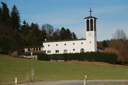 Ein weißes Kirchengebäude mit Turm, Uhr und Kreuz steht in einer ländlichen Gegend, umgeben von grünen Bäumen und Büschen.