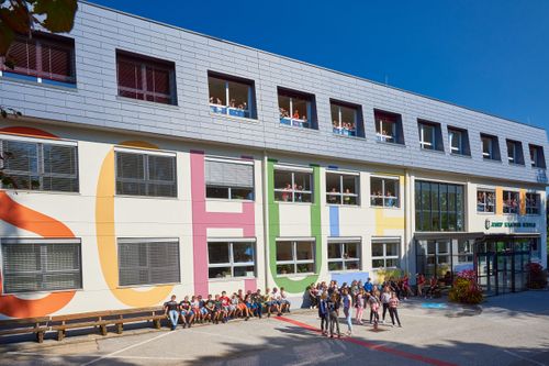 Ein Schulgebäude mit bunten Buchstaben an der Wand, Schüler sitzen und stehen draußen, einige winken aus den Fenstern, und ein klarer blauer Himmel im Hintergrund.