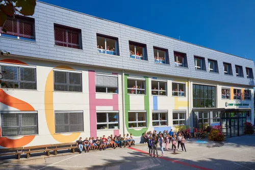 Ein Schulgebäude mit bunten Buchstaben an der Wand, Schüler sitzen und stehen draußen, einige winken aus den Fenstern, und ein klarer blauer Himmel im Hintergrund.